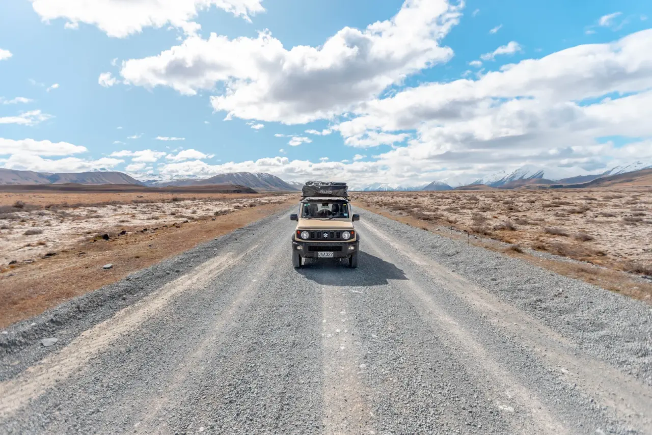 Tan Suzuki Jimny with rooftop tent on gravel road with snow-capped mountains, South Island